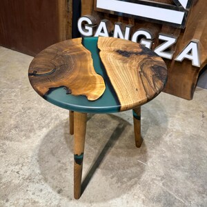 May include: A round wooden side table with a teal epoxy river design and three wooden legs. The table is on a concrete floor.