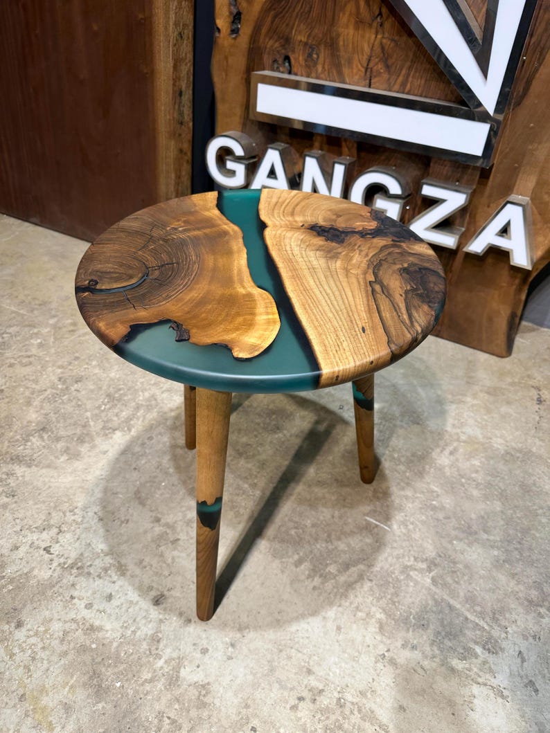 May include: A round wooden side table with a teal epoxy river design and three wooden legs. The table is on a concrete floor.