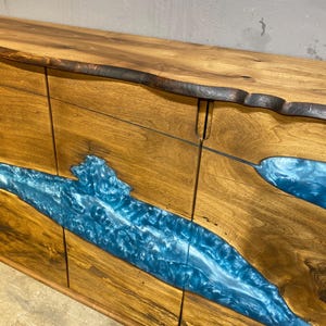 May include: A wooden sideboard with a live edge top and a blue resin river design. The cabinet doors are made of wood with a natural grain, and the resin flows across the front. The wood has a warm, natural tone.