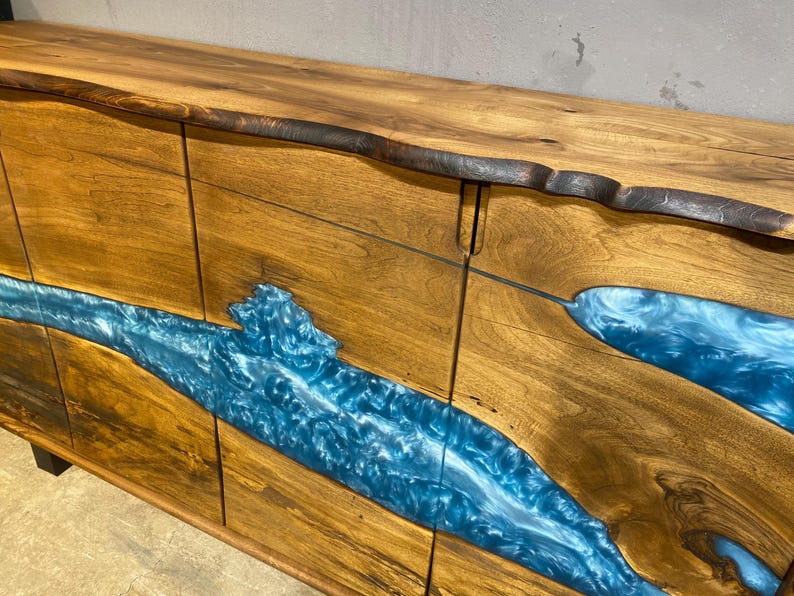 May include: A wooden sideboard with a live edge top and a blue resin river design. The cabinet doors are made of wood with a natural grain, and the resin flows across the front. The wood has a warm, natural tone.