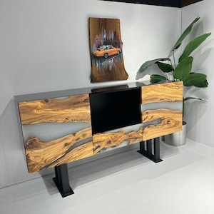 May include: A modern wooden cabinet with a black and white epoxy resin design. The cabinet has two doors and a shelf. It is standing on black metal legs. A painting of a car in a city scene hangs on the wall behind the cabinet.