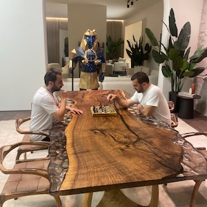 May include: A large live-edge wooden dining table with a central resin river inlay, featuring a natural wood grain pattern and dark edges. The table is supported by a curved wooden base and is surrounded by matching wooden chairs with brown cushions.