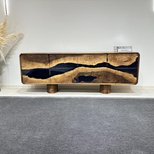 May include: A wooden sideboard with black resin inlay. The sideboard has four doors and sits on four legs. The sideboard has a book on top with the text "TOM FORD" and "CHANEL" visible.