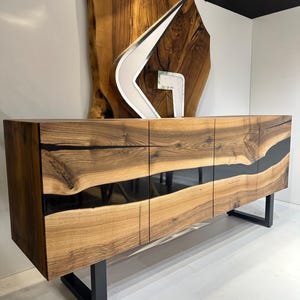 May include: A wooden sideboard with a natural wood grain finish and black resin accents. The cabinet has four doors and sits on black metal legs. A decorative sculpture and a large wooden slab are in the background.