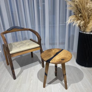 Puede incluir: Una silla de madera con respaldo curvo y asiento acolchado, junto a una pequeña mesa redonda de madera con incrustaciones de resina negra. La mesa tiene tres patas.