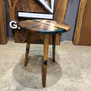 May include: A small round wooden side table with three legs. The table top is made of wood with a blue and black epoxy resin inlay.
