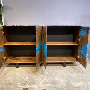 May include: Wooden cabinet with two doors and two shelves. The cabinet is made of brown wood with blue resin accents. The doors are open, revealing the shelves inside. The cabinet is on a gray floor.