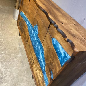 May include: A wooden cabinet with a natural edge design. The cabinet features a river of blue resin flowing across the front. The wood has a warm brown tone, and the resin has a glossy finish.