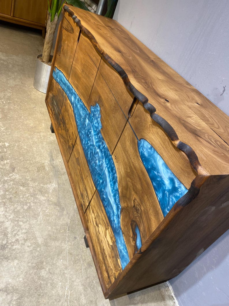 May include: A wooden cabinet with a natural edge design. The cabinet features a river of blue resin flowing across the front. The wood has a warm brown tone, and the resin has a glossy finish.