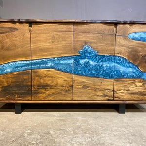 May include: A wooden cabinet with four doors, featuring a river of blue resin flowing through the natural wood grain. The cabinet is rectangular, with a dark brown frame and black legs. The wood has a warm, natural tone.