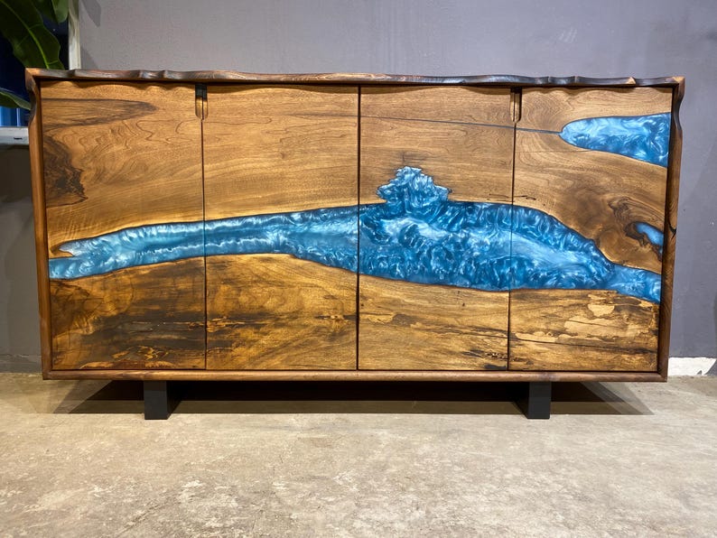 May include: A wooden cabinet with four doors, featuring a river of blue resin flowing through the natural wood grain. The cabinet is rectangular, with a dark brown frame and black legs. The wood has a warm, natural tone.