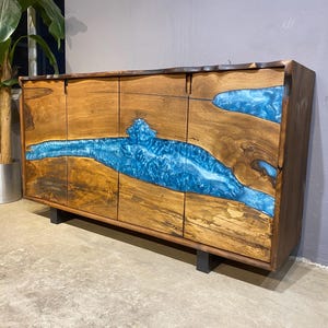 May include: A wooden sideboard with a unique design. The cabinet features a natural wood grain finish with sections of vibrant blue resin, creating a river-like effect. It has four doors and sits on black metal legs. A modern and stylish furniture piece.