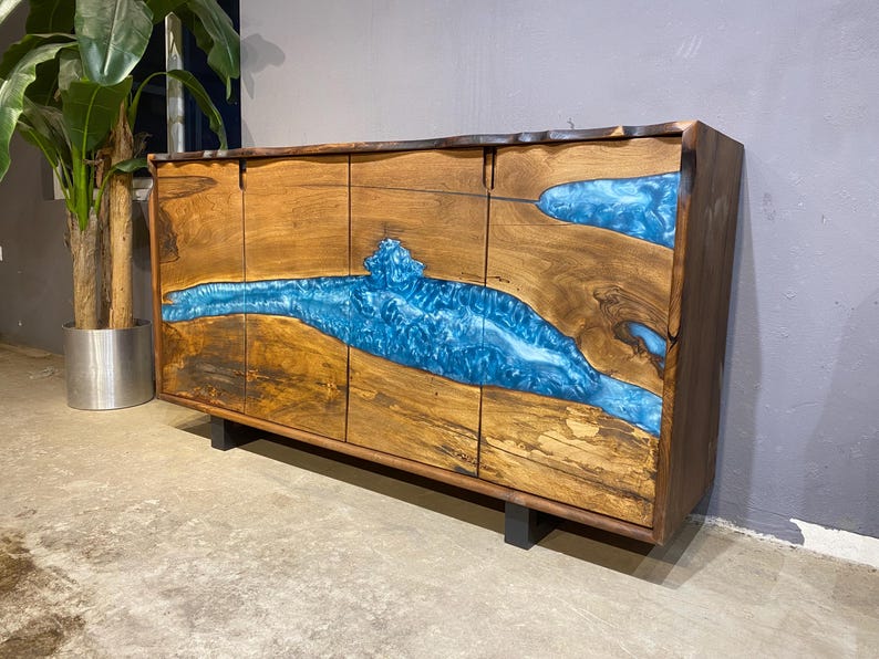 May include: A wooden sideboard with a unique design. The cabinet features a natural wood grain finish with sections of vibrant blue resin, creating a river-like effect. It has four doors and sits on black metal legs. A modern and stylish furniture piece.