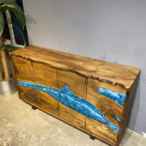 May include: A wooden cabinet with a natural edge design. The cabinet features a blue resin river design across the front. The wood has a warm brown tone, and the cabinet sits on black legs. A modern piece of furniture.