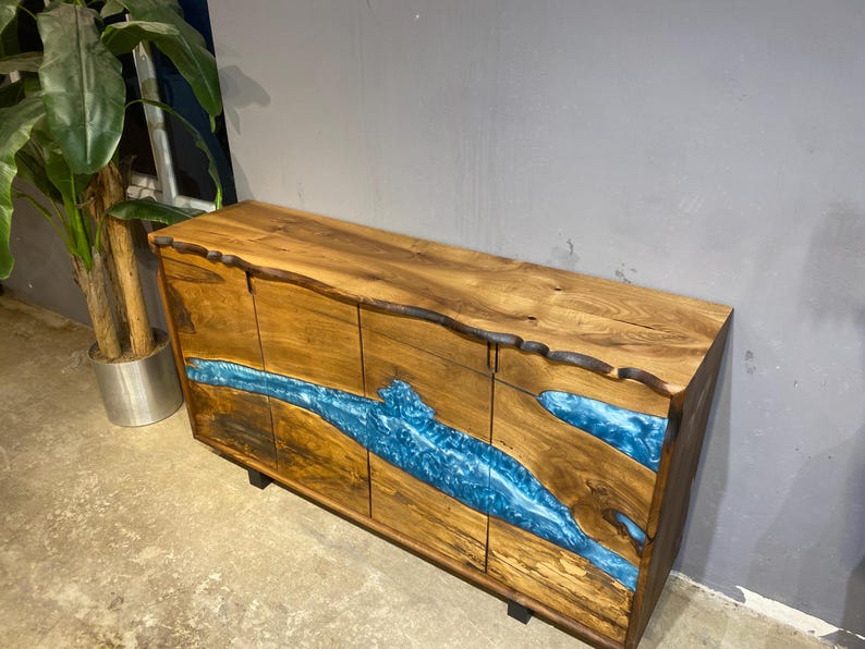 May include: A wooden cabinet with a natural edge design. The cabinet features a blue resin river design across the front. The wood has a warm brown tone, and the cabinet sits on black legs. A modern piece of furniture.