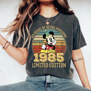 May include: Dark gray t-shirt featuring a vintage design with Mickey Mouse wearing a party hat. The shirt includes the text "40 YEARS OF BEING AWESOME" and "1985 LIMITED EDITION" in a retro style, with a colorful sunset background.