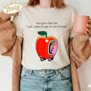 Könnte beinhalten: Ein elfenbeinfarbenes T-Shirt mit dem schwarzen Text "You got a fast car. I got a plan to get us out of here." und einer Grafik eines roten Apfels in Form eines Autos mit einem Wurm darin, getragen unter einer braunen Strickjacke mit einem rosa Becher.