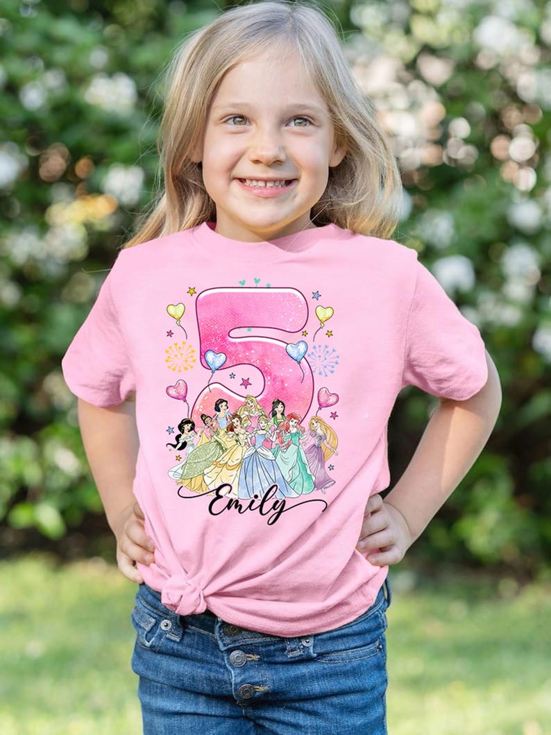 A young girl wearing a pink t-shirt with a Disney princess design and the number 5, smiling at the camera.