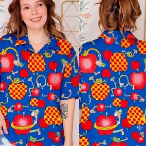 Könnte beinhalten: Ein kurzärmeliges Hemd mit Knopfleiste in leuchtendem Blau, das mit einem durchgehenden Muster aus roten Äpfeln, gelben Äpfeln mit roten Punkten und kleinen grünen Würmern bedruckt ist. Das Hemd hat einen klassischen Kragen und eine Knopfleiste.