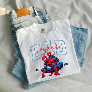 Può includere: Una maglietta bianca con una grafica di Spider-Man e le parole "Super Dad" in blu e rosso. La maglietta è piegata sopra un paio di jeans blu chiaro.
