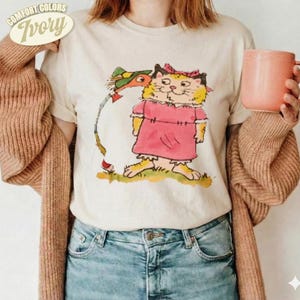 Könnte beinhalten: Ein elfenbeinfarbenes T-Shirt mit einer grafischen Illustration einer gelben Katze in einem rosa Kleid mit rosa Schleife, die neben einem grünen Vogel steht. Das Shirt wird unter einer beigen Strickjacke zu blauen Jeans getragen und hält eine pfirsichfarbene Tasse.