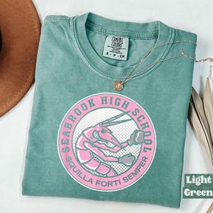 May include: A light green t-shirt with a circular graphic featuring a pink shrimp and the words "SEABROOK HIGH SCHOOL" and "SQUILLA FORTI SEMPER". A silver necklace with a small pendant rests on the shirt. The shirt is labeled "Comfort Colors".