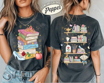 May include: Dark gray t-shirt with a colorful graphic of stacked books, a tiara, and fairy tale elements. The back of the shirt features a bookshelf design with books and other fairy tale items. The text "COMFORT COLORS Pepper" is on the top.