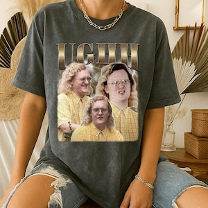 May include: Dark grey t-shirt with a graphic of a man wearing glasses and a yellow plaid shirt. The word "LIGHT" is printed above the image in a gold, textured font. The shirt has short sleeves.