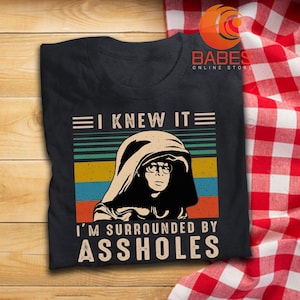 May include: Black t-shirt with a graphic design featuring a person in a hood and glasses, with the text "I KNEW IT" above and "I'M SURROUNDED BY ASSHOLES" below. The design has a retro color scheme.