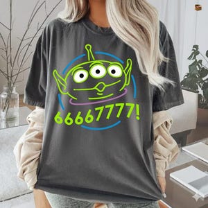 May include: Dark gray t-shirt featuring a cartoon alien with three eyes, green outline, and the text "66667777!" in green and blue. The alien is inside a blue circle with a purple line below the alien's mouth.