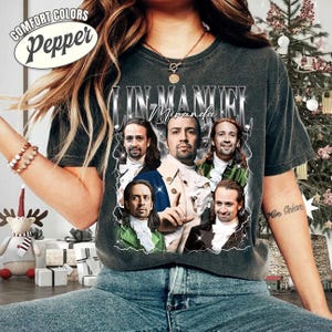 May include: Dark gray t-shirt featuring a graphic of Lin-Manuel Miranda with the text "Lin-Manuel Miranda". The shirt has a distressed look and the words "Comfort Colors Pepper" are printed on the top left.