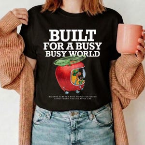 Könnte beinhalten: Ein schwarzes Kurzarm-T-Shirt mit dem weißen Text BUILT FOR A BUSY BUSY WORLD über einer Grafik eines roten apfelförmigen Autos mit einem grünen Blatt oben und einer Wurmfigur im Inneren. Kleiner Text darunter lautet RICHARD SCARRY'S BUSY WORLD.