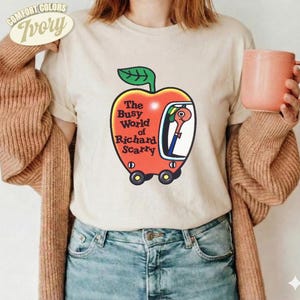 Könnte beinhalten: Ein elfenbeinfarbenes Kurzarm-T-Shirt mit einer Grafik eines roten apfelförmigen Autos mit einem grünen Blatt oben. Im Auto befindet sich ein Wurm. Der Text lautet The Busy World of Richard Scarry. Getragen mit einer braunen Strickjacke.