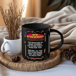 以下が含まれることがあります： 「NEW ACHIEVEMENT!」の文字とピクセルフォントのテキストが入った黒いセラミックマグカップ。マグカップには黒いハンドルが付いており、白いマグカップと装飾要素のある織りのトレイの上に置かれています。