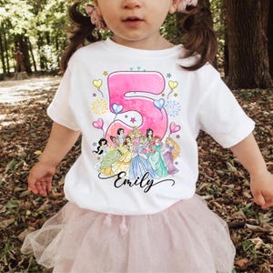A young girl wearing a white t-shirt with a Disney princess design and the number 5, along with the name "Emilly".