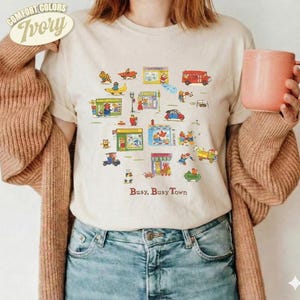 Könnte beinhalten: Ein elfenbeinfarbenes Kurzarm-T-Shirt mit einem bunten Grafikdruck verschiedener Gebäude, Fahrzeuge und Figuren sowie dem Text "Busy, Busy Town" unter der Illustration. Das T-Shirt wird mit einer braunen Strickjacke und blauen Jeans getragen.