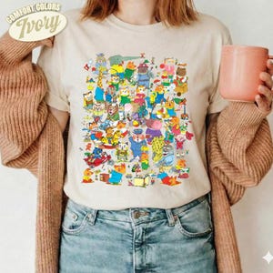 Könnte beinhalten: Ein elfenbeinfarbenes Kurzarm-T-Shirt mit einem großen, bunten Grafikdruck zahlreicher Zeichentricktiere bei verschiedenen Aktivitäten. Das T-Shirt wird mit einer braunen Langarmstrickjacke und einer blauen Jeans getragen.