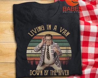 May include: Black t-shirt with a graphic featuring a man in a plaid jacket and tie, with the text "LIVING IN A VAN DOWN BY THE RIVER" in a retro style. The shirt has a crew neck.