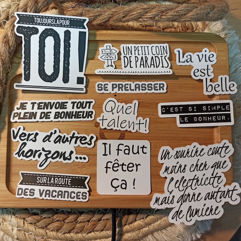 May include: A collection of nine white stickers with black text and illustrations. The stickers feature motivational phrases in French, such as "Toi!" (You!), "Un petit coin de paradis" (A little corner of paradise), "Se prelasser" (To relax), "Quel talent!" (What talent!), "Il faut f&ecirc;ter &ccedil;a!" (We must celebrate this!), "Je t'envoie tout plein de bonheur" (I send you lots of happiness), "Vers d'autres horizons..." (Towards other horizons...), "C'est si simple le bonheur" (It's so simple, happiness), and "Un sourire co&ucirc;te moins cher que l'&eacute;lectricit&eacute; mais donne autant de lumi&egrave;re" (A smile costs less than electricity but gives as much light).