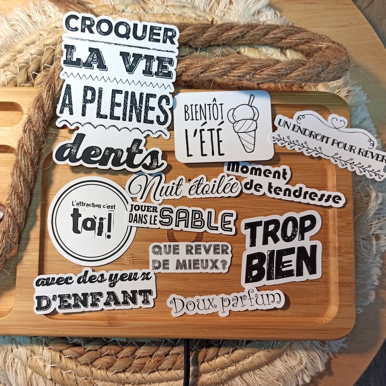 May include: A collection of black and white stickers with French phrases. The stickers are on a wooden surface. The phrases include "Croquer la vie a pleines dents", "Bientot l'ete", "Un endroit pour rever", "Moment nuit etoile de tendresse", "L'attraction c'est toi", "Jouer dans le sable", "Que rever de mieux", "Trop bien", "Avec des yeux d'enfant", and "Doux parfum".
