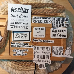 May include: A collection of white stickers with black text on a wooden surface. The stickers feature French phrases such as "Des c&acirc;lins tout doux" and "La magie d'un instant pr&eacute;cieux".