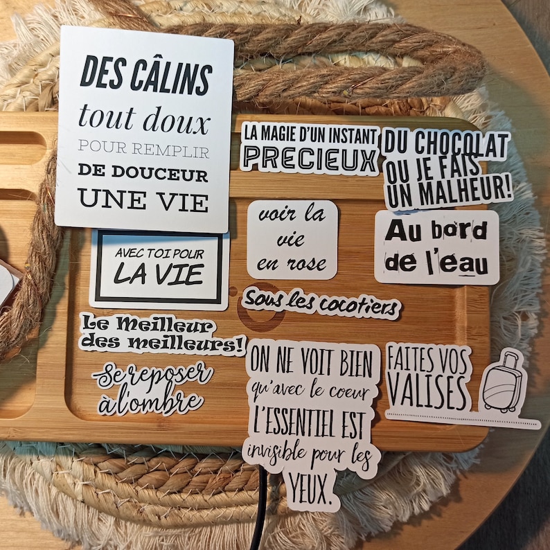 May include: A collection of white stickers with black text on a wooden surface. The stickers feature French phrases such as "Des c&acirc;lins tout doux" and "La magie d'un instant pr&eacute;cieux".