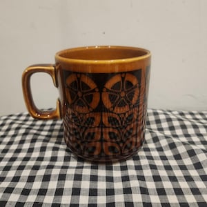 May include: A ceramic mug featuring a glossy amber-brown glaze with a dark brown geometric pattern. The design consists of repeating circular motifs and stylised leaf shapes arranged in vertical columns around the exterior of the cylindrical vessel.