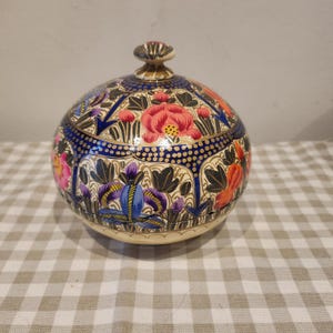 May include: A spherical lidded container featuring a hand-painted floral pattern with vibrant red, orange, purple, and pink blossoms against a cream background. The design includes intricate dark blue geometric borders with gold dot accents.
