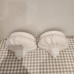May include: Two white ceramic wall sconces with detailed floral and scrollwork. The sconces have a rounded top and a tapered base. They are displayed on a checkered fabric. Each sconce is approximately 20 cm wide.