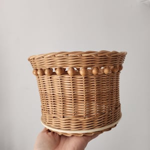 May include: A light brown woven wicker basket with a cylindrical shape and a flat base. The upper section features a decorative row of small, round wooden beads integrated into the weave, creating a textured border around the top rim.
