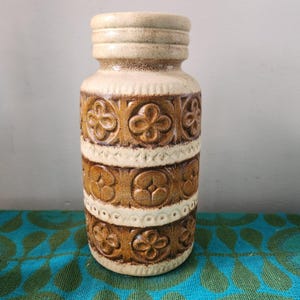 May include: A vintage ceramic vase in beige and brown. The cylindrical vase has a textured surface and floral patterns. It is approximately 25 cm tall. A decorative piece for the home.