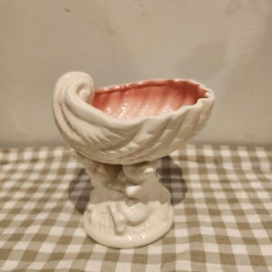 May include: A white ceramic seashell-shaped bowl with a pink interior. The bowl is supported by a pedestal base with a sculpted design. The bowl has a textured, ridged exterior and is likely used for decorative purposes.