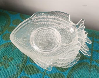 May include: Clear glass fish-shaped coasters. The coasters are stacked, showing the fish scales and fin details. Each coaster has a circular indentation in the center. The fish design is a decorative element for home decor.