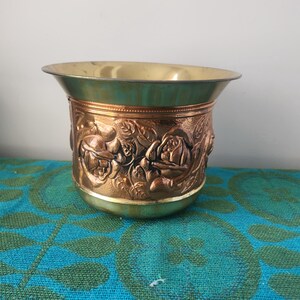 May include: A brass and copper planter with a detailed floral design. The planter has a wide, flared rim and a decorative band featuring embossed roses. The interior is a lighter brass colour, and the exterior has a mix of copper and brass tones.
